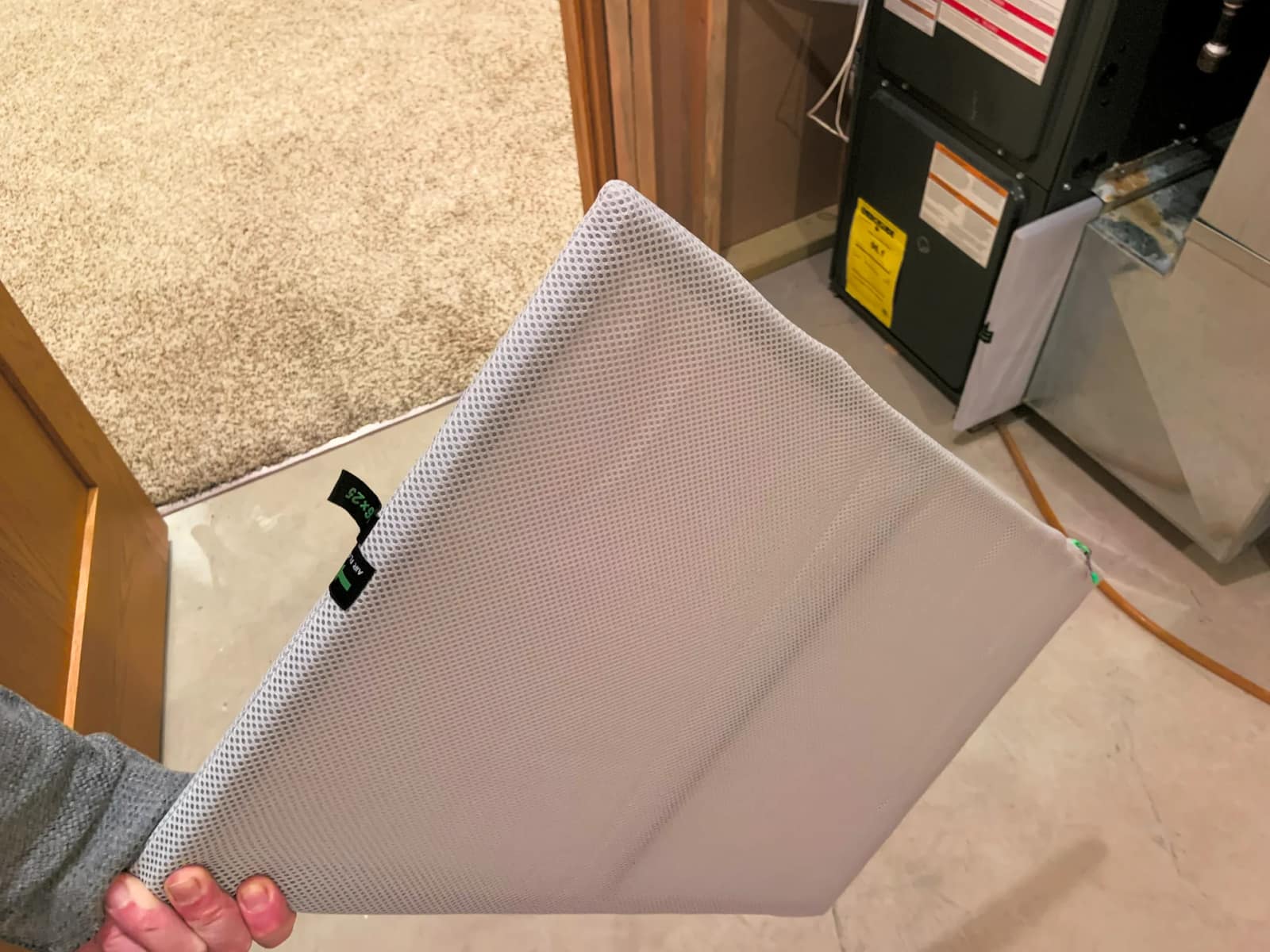 A hand holding a gray mesh furnace air filter in a basement, with the furnace and its filter slot behind it.