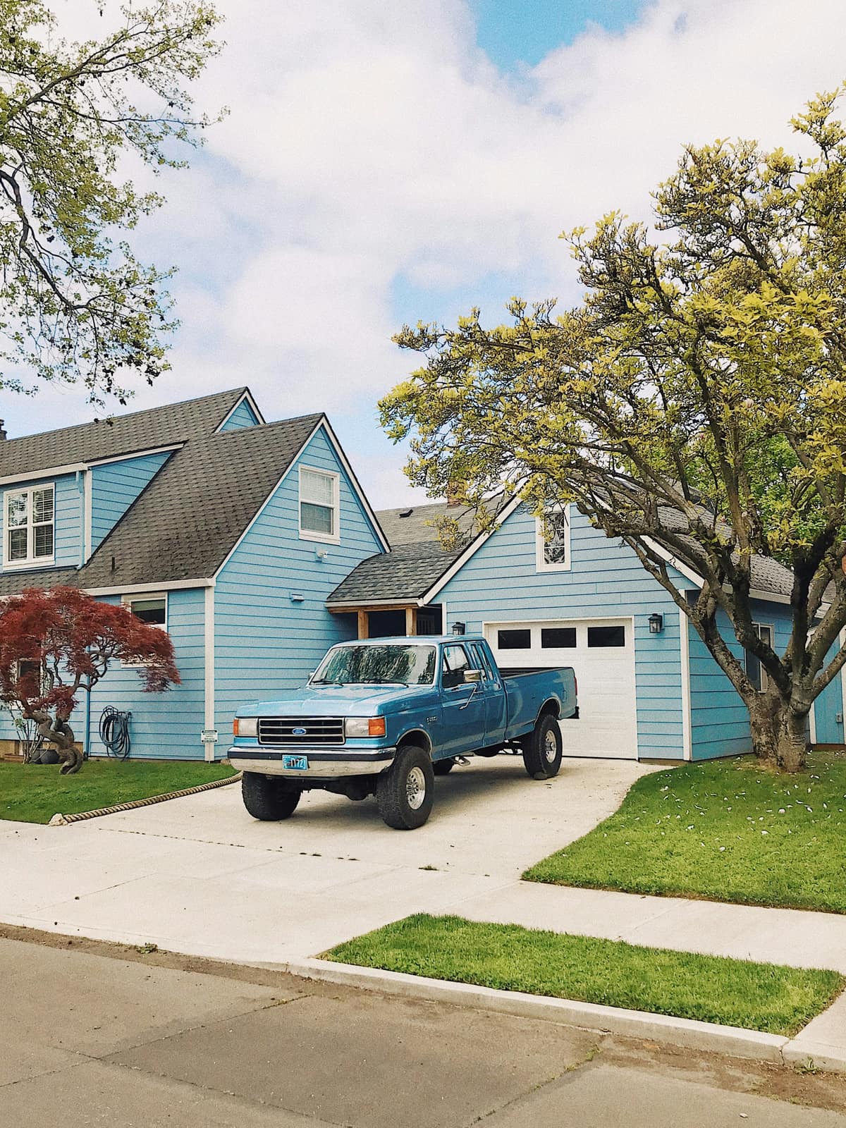 A blue Ford pickup truck parked in the driveway of a blue suburban house with a tree and lawn.