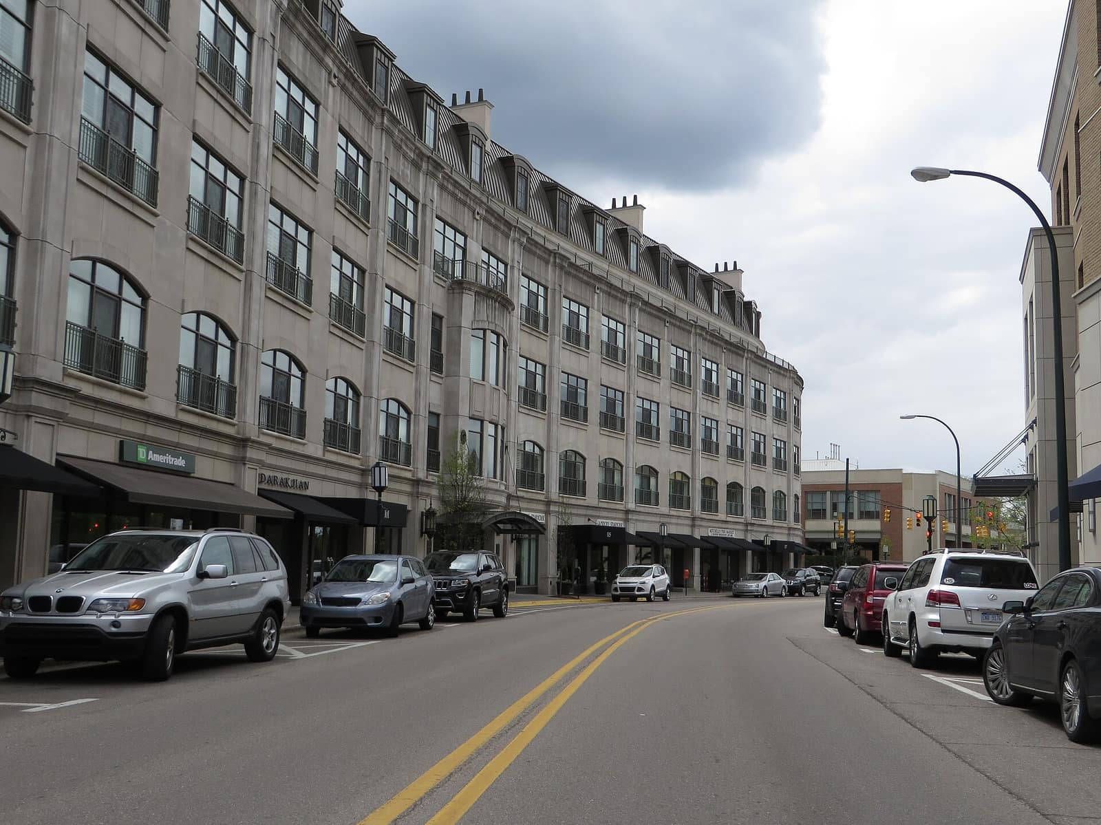 A downtown Birmingham, Michigan street lined with parked cars and a large stone-faced mixed-use building under a cloudy sky.