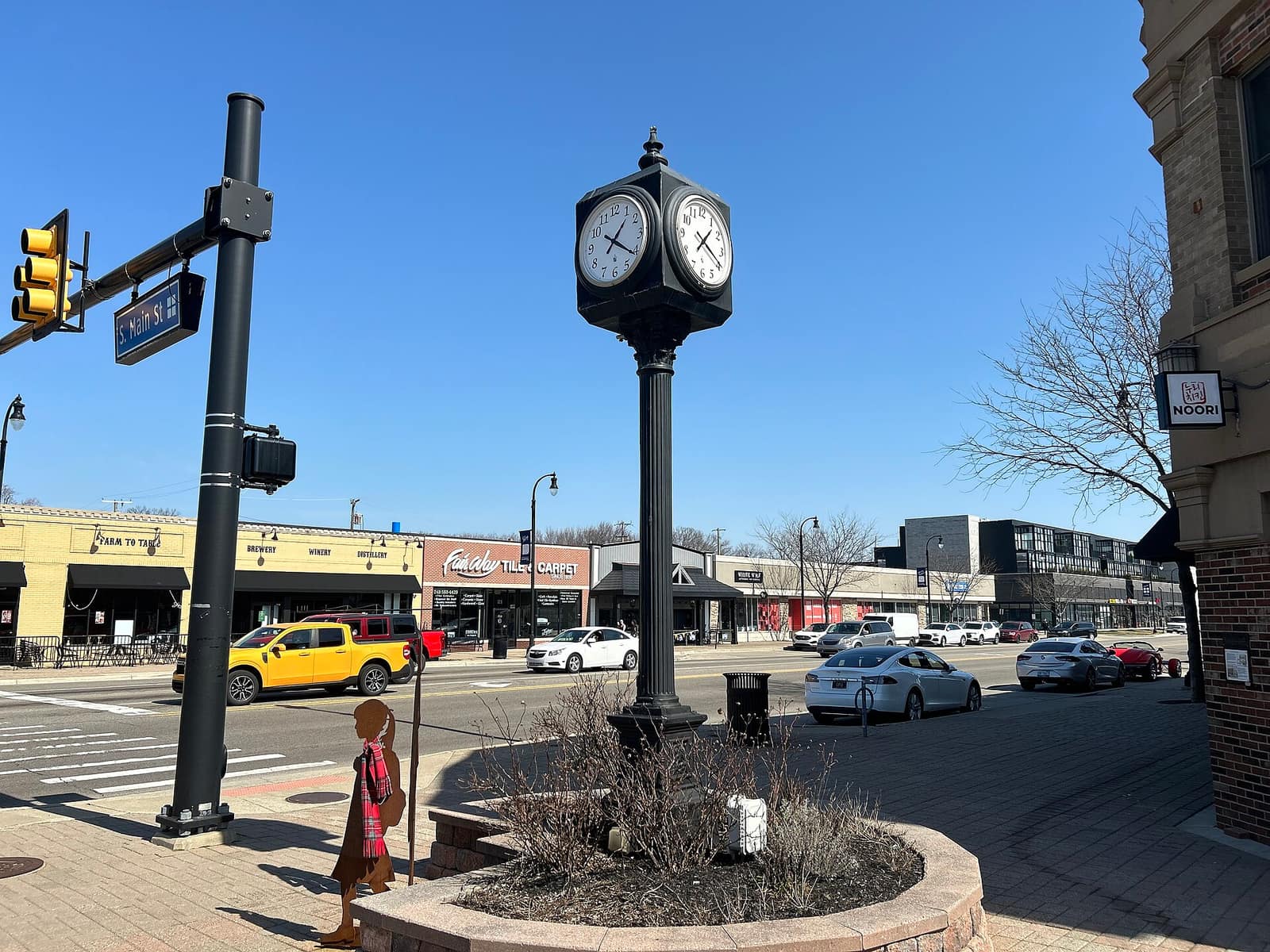 The street clock at Main Street and 14 Mile in downtown Clawson, Michigan, with storefronts and parked cars along the road.