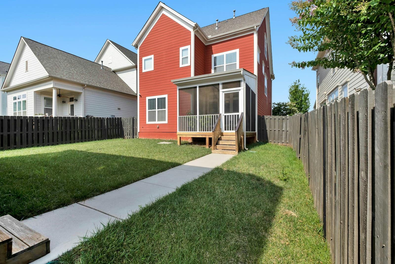 A narrow fenced backyard lawn with a concrete walk leading to a red house with a screened porch, wooden fences on both sides.