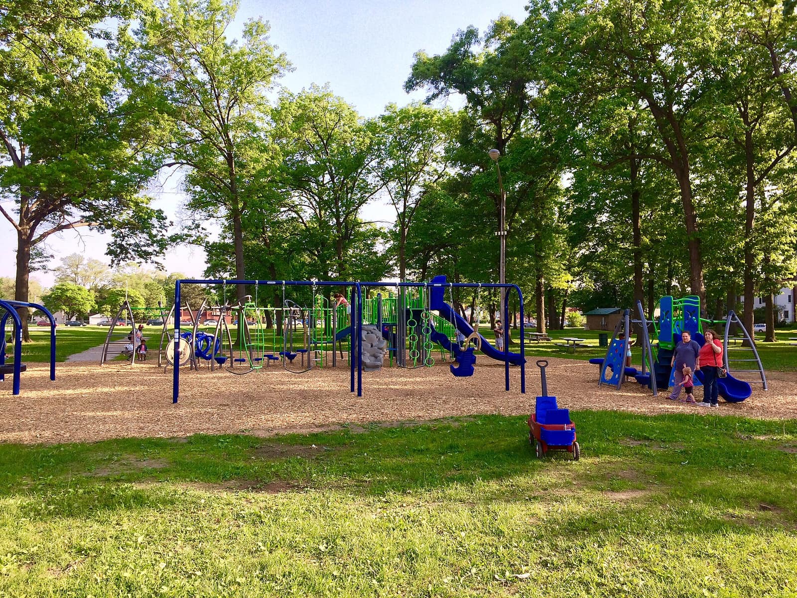 A playground with blue and green equipment on a woodchip surface under tall shade trees at Garbutt Park in Ferndale, Michigan, on a spring evening.