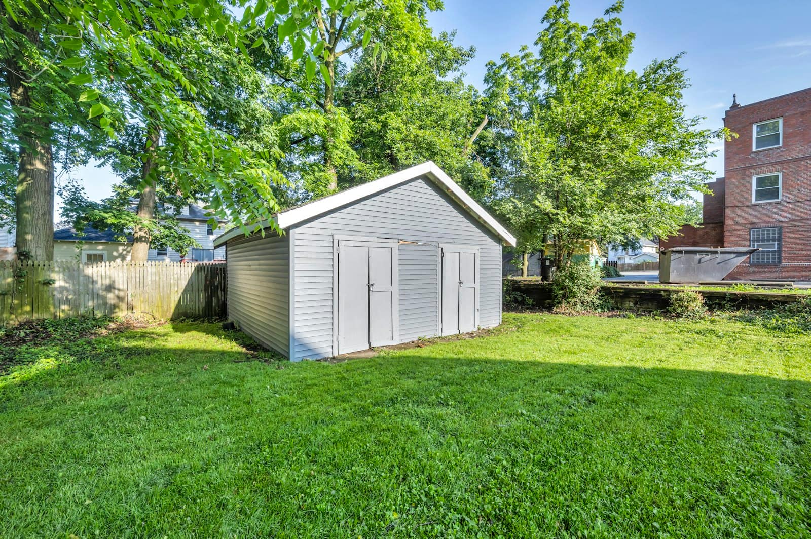 A small grassy backyard with a grey outbuilding, a wooden fence and older houses and trees behind it.