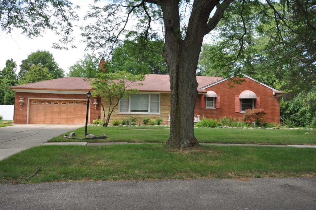 A red and tan brick ranch house with an attached garage behind a mown front lawn and a large tree.