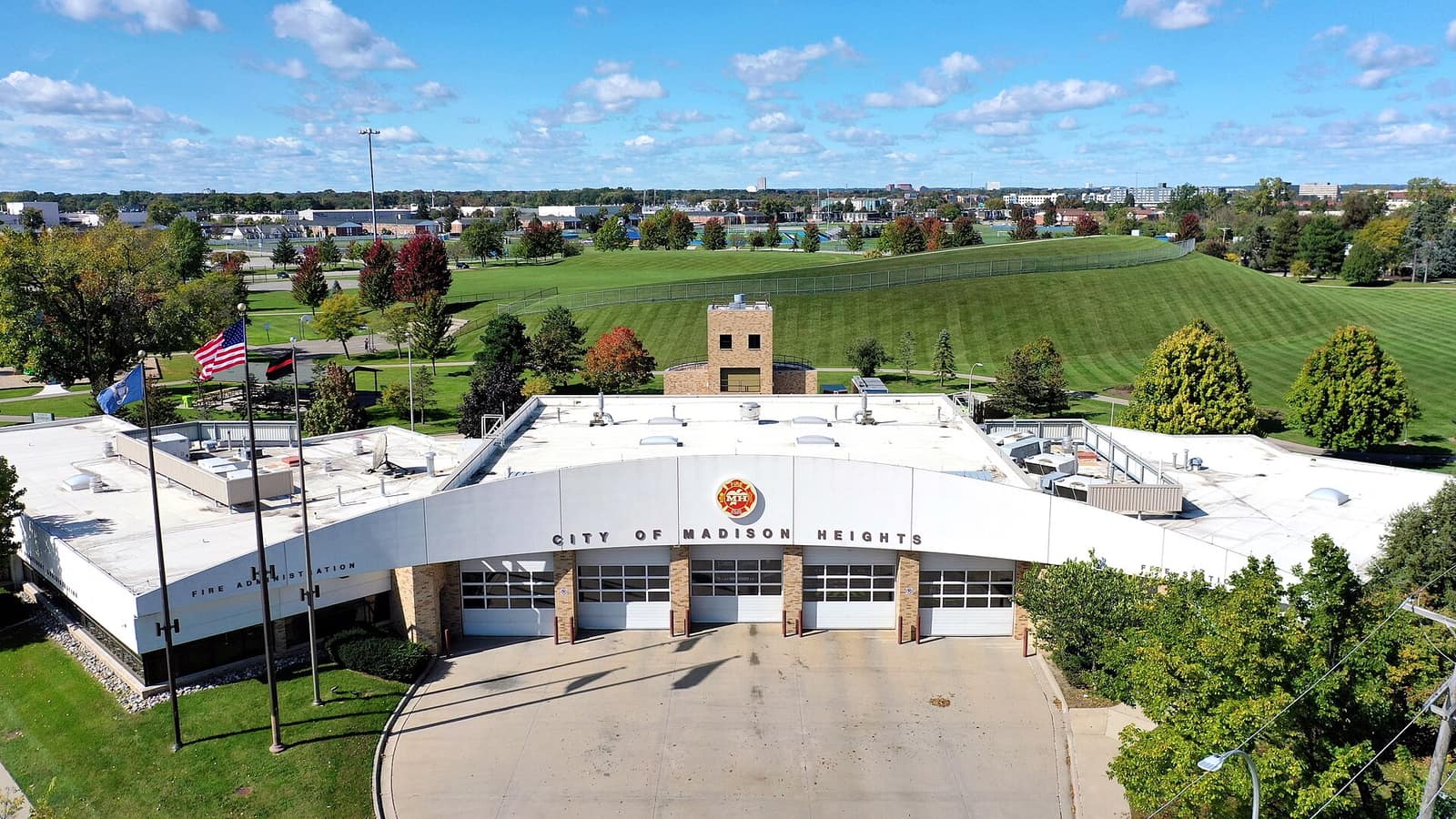 An aerial view of the white arched front of Madison Heights Fire Station 1 with its apron, flags and a grass park behind.