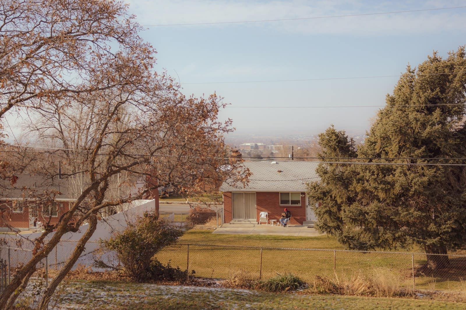A brick ranch house across an open, fenced backyard lawn in late autumn, with bare trees.