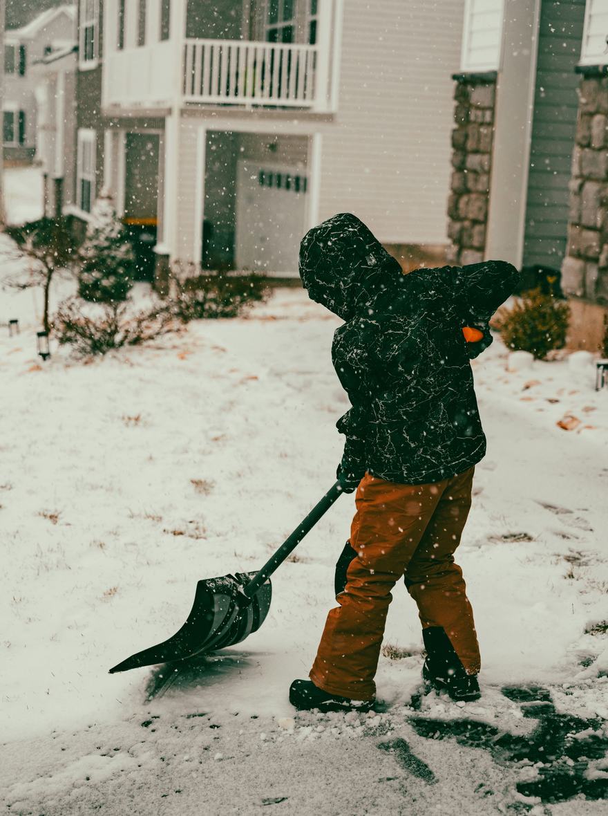 A kid pushing a snow shovel down a snowy front walk.